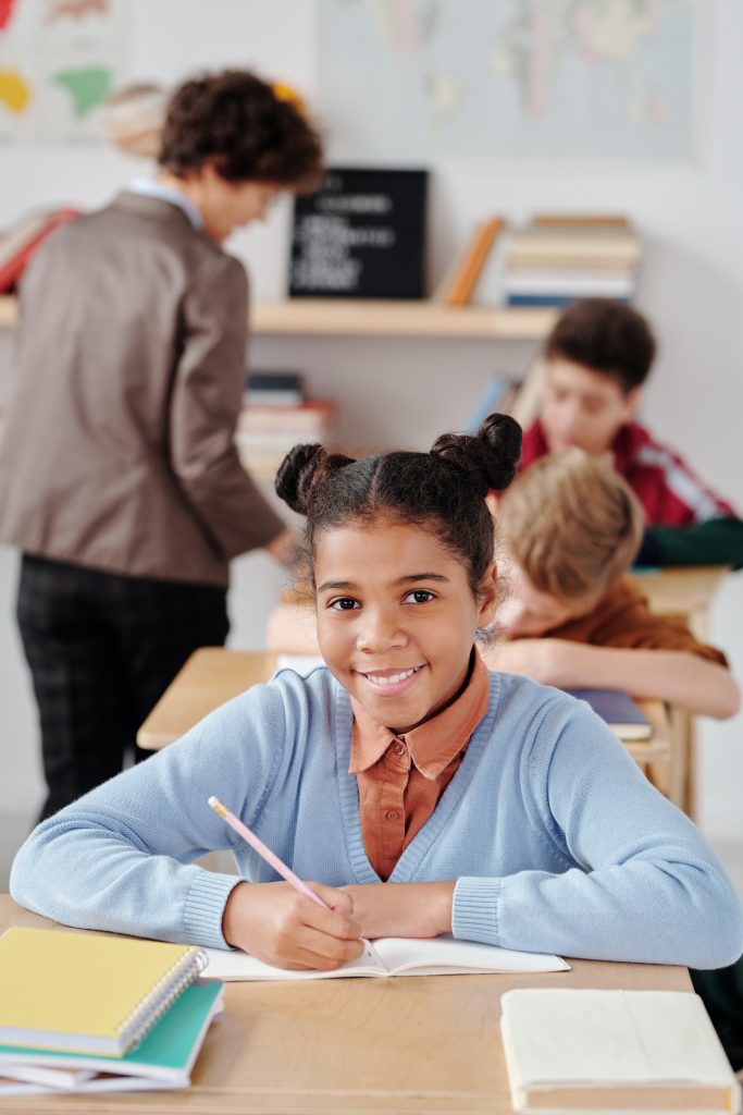 Young girl in long sleeve sweater sitting at desk with pencil in hand smiling while teacher helps students in the background