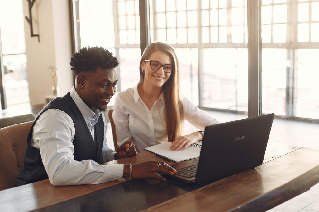 Two co-workers sitting at a table with big windows in the background, having a conversation while looking at a laptop