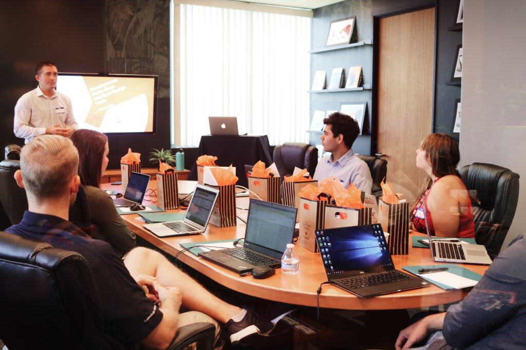 Group of people sitting around a meetin gtable. One man stands at the head of the table presenting information