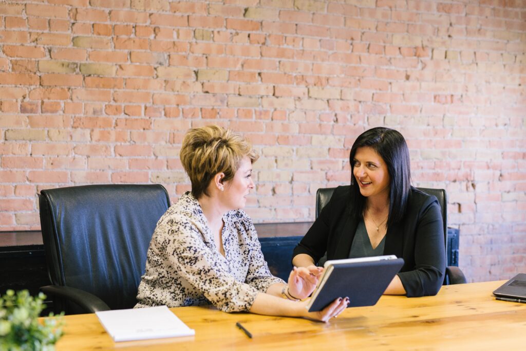 Two women sitting side by side at table talking while looking at computer screen