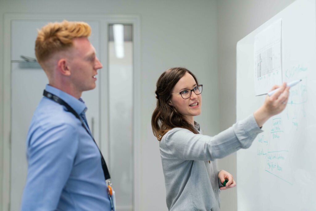 Two people are working together at the whiteboard. A woman in a gray shirt holds a pen explaining something to the man who stands behind her in a blue shirt.