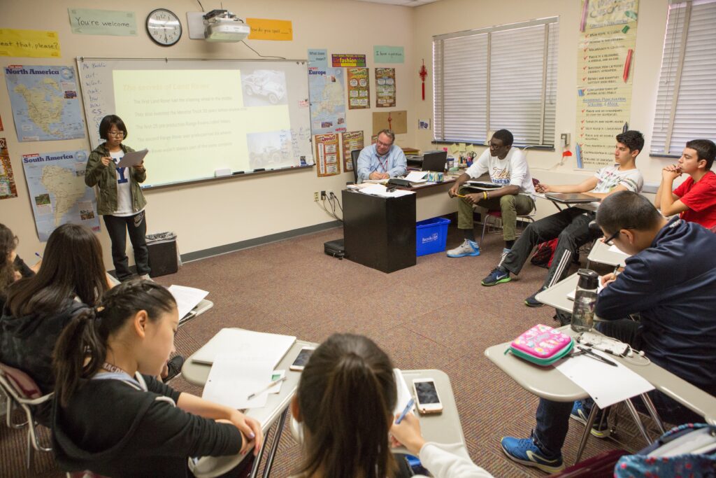 image of classroom with students seated at desks in a circle and one student at the front of the room presenting