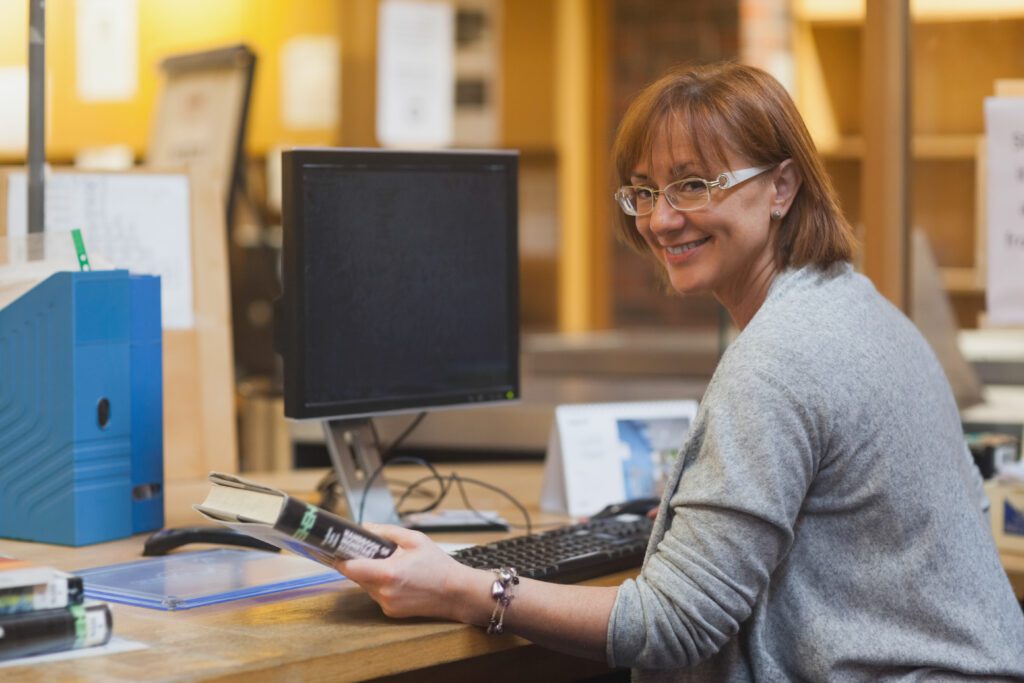 Smiling female librarian holding a book sitting behind the desk looking at camera
