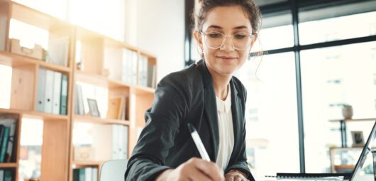 Woman writing in notebook while sitting in front of a computer